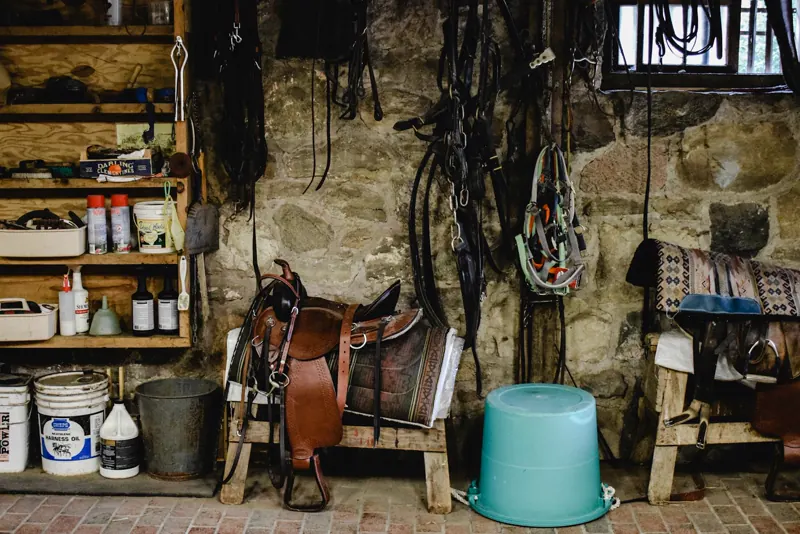 Sala de arreios com selas, rédeas e equipamentos equestres organizados em armários de madeira e paredes de pedra